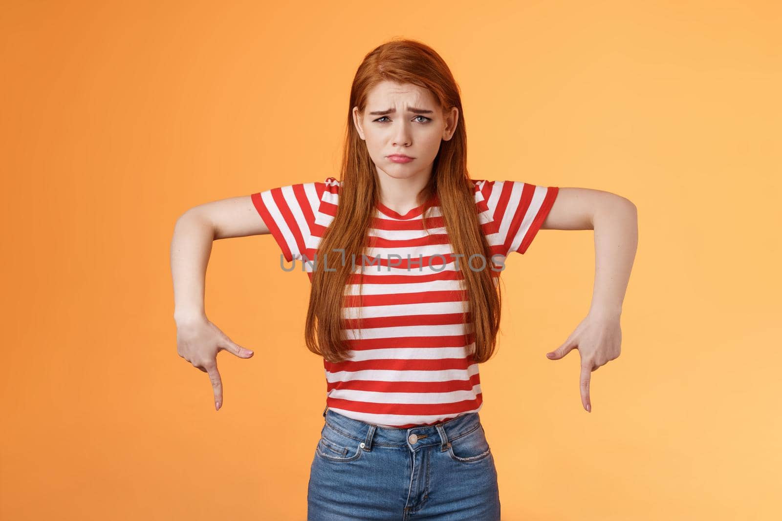 Cute timid hesitant ginger female taking tough decision feel pressured upset, frowning, pulling gloomy unhappy face, pointing down disappointed, uneasy taking decision, orange background.