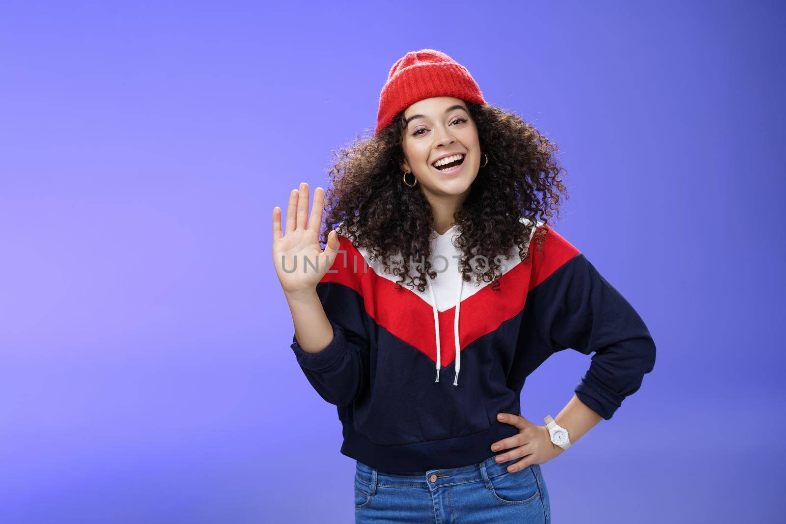 Hey nice to meet you. Charming female ski couch in cute red beanie with curly hair waving hello with palm and smiling broadly greeting newbies as teaching winter sports over blue background. Copy space