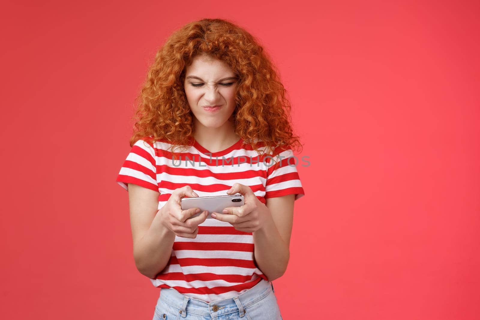 Daring enthusiastic addicted geeky good-looking redhead curly girl hate losing battles smartphone game wrinkle nose grimacing upset eager win tap fast telephone screen look display, red background by Benzoix