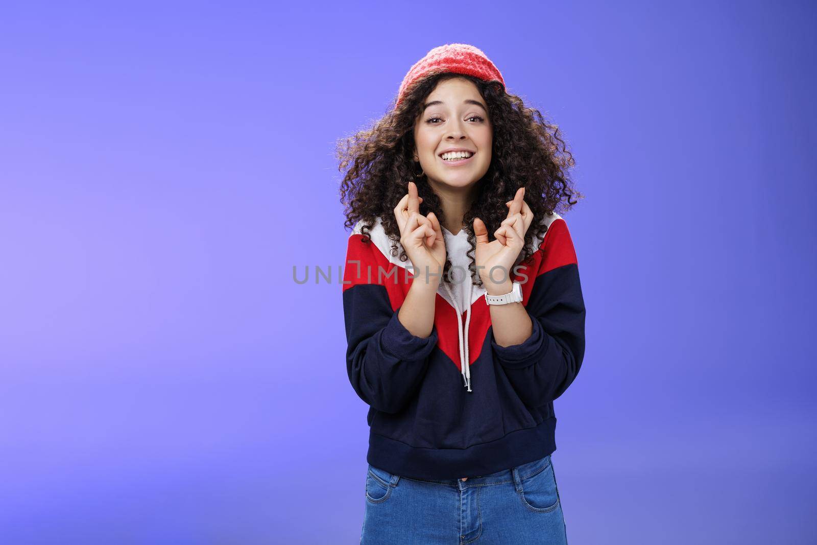 Optimistic and hopeful girl having faith in desire come true, standing with crossed fingers for good luck having wish anticipating miracle, smiling broadly at camera as wearing warm stylish red beanie.