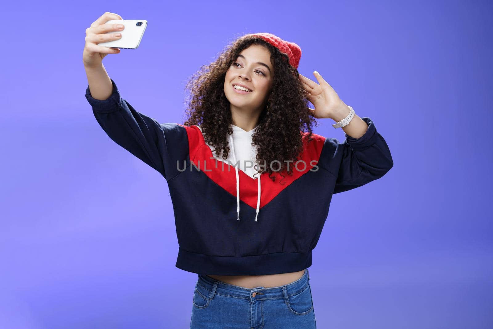Cute girl making selfie to memorize trip standing in warm winter beanie and sweatshirt posing as pulling hand with smartphone and smiling at device screen to take pretty shot and post internet. Technology and lifestyle concept