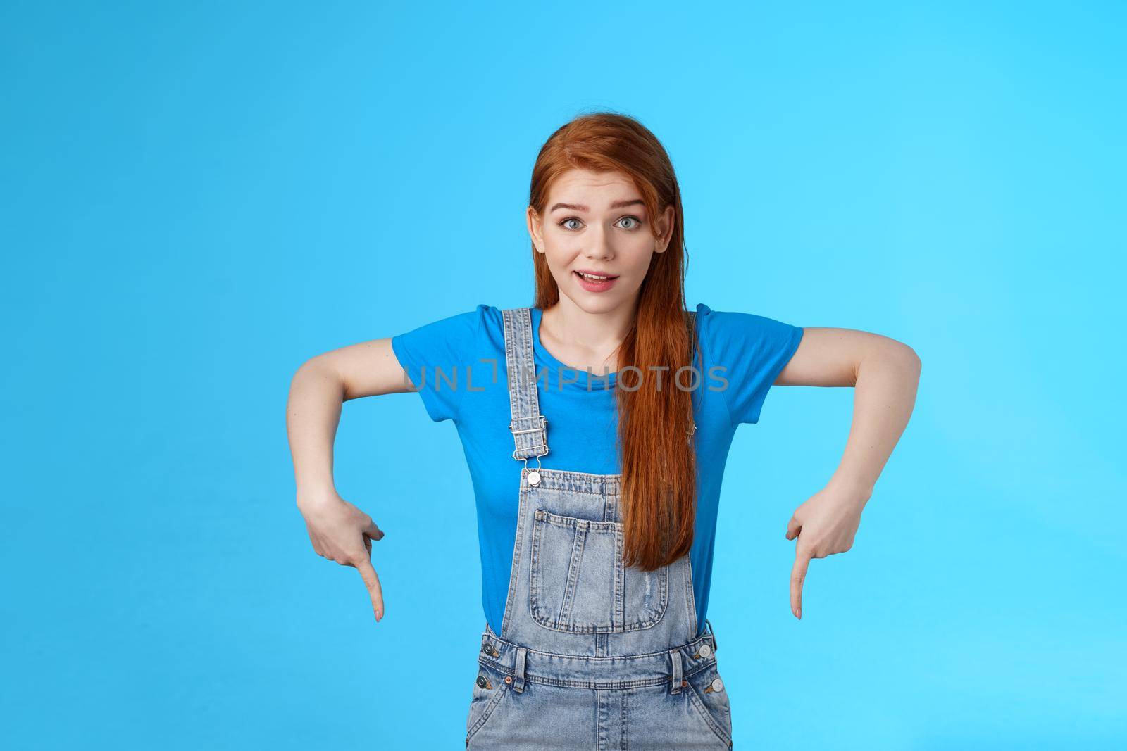 Curious tender lovely ginger caucasian girl, pointing down questioned, check out interesting promo, point bottom down copy space, look camera intrigued, suggest good product online shop link.