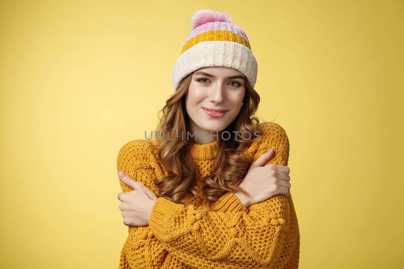 Attractive feminine tender young woman feel cozy, embracing herself smiling delighted cute, wearing comfortable warm sweater trendy hat cuddling, dreaming boyfriend hugs, stand yellow background.