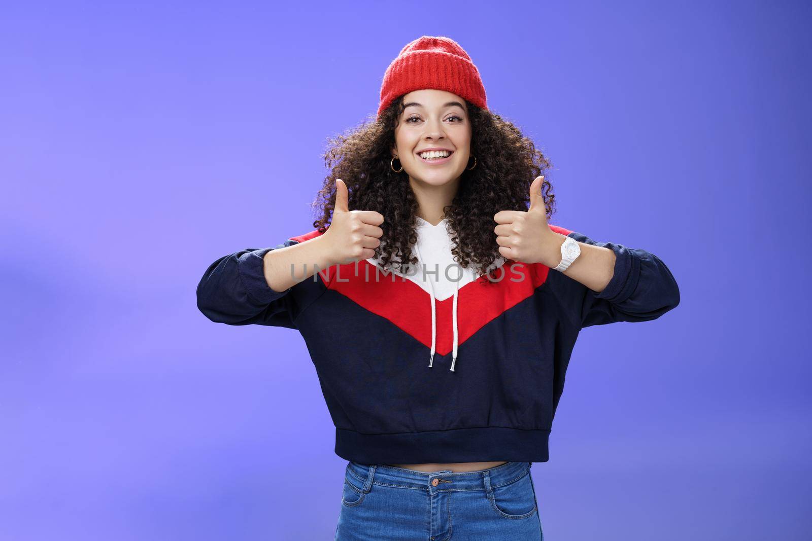 Girl totally agrees recommends cool place showing thumbs-up and smiling excited with happy grin standing upbeat and delighted against blue background in warm beanie and sweatshirt by Benzoix