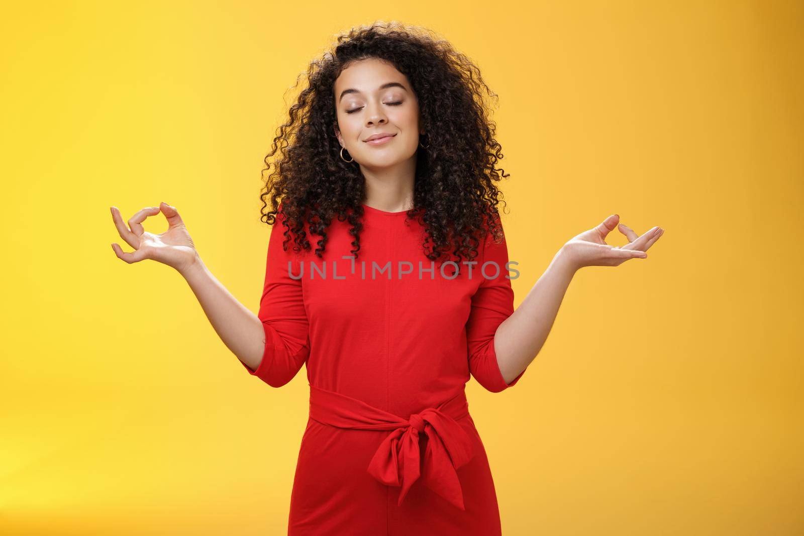 Charming dreamy and happy peaceful woman in red dress with curly hair standing in lotus pose with mudra orbs or zen gesture and closed eyes, meditation relieving stress, feeling unbothered. Body language, buddhism and people concept