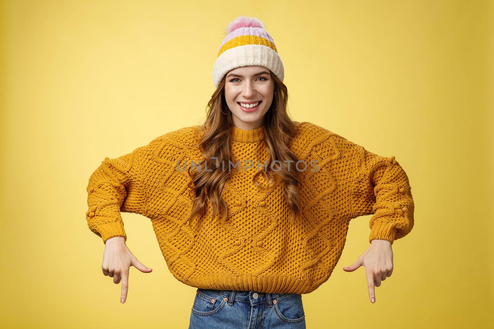 Lifestyle. Friendly-looking helpful good-looking 20s college student girl wearing hat warm sweater smiling pleased showing you awesome promotion pointing index fingers down recommend bottom advertisement.