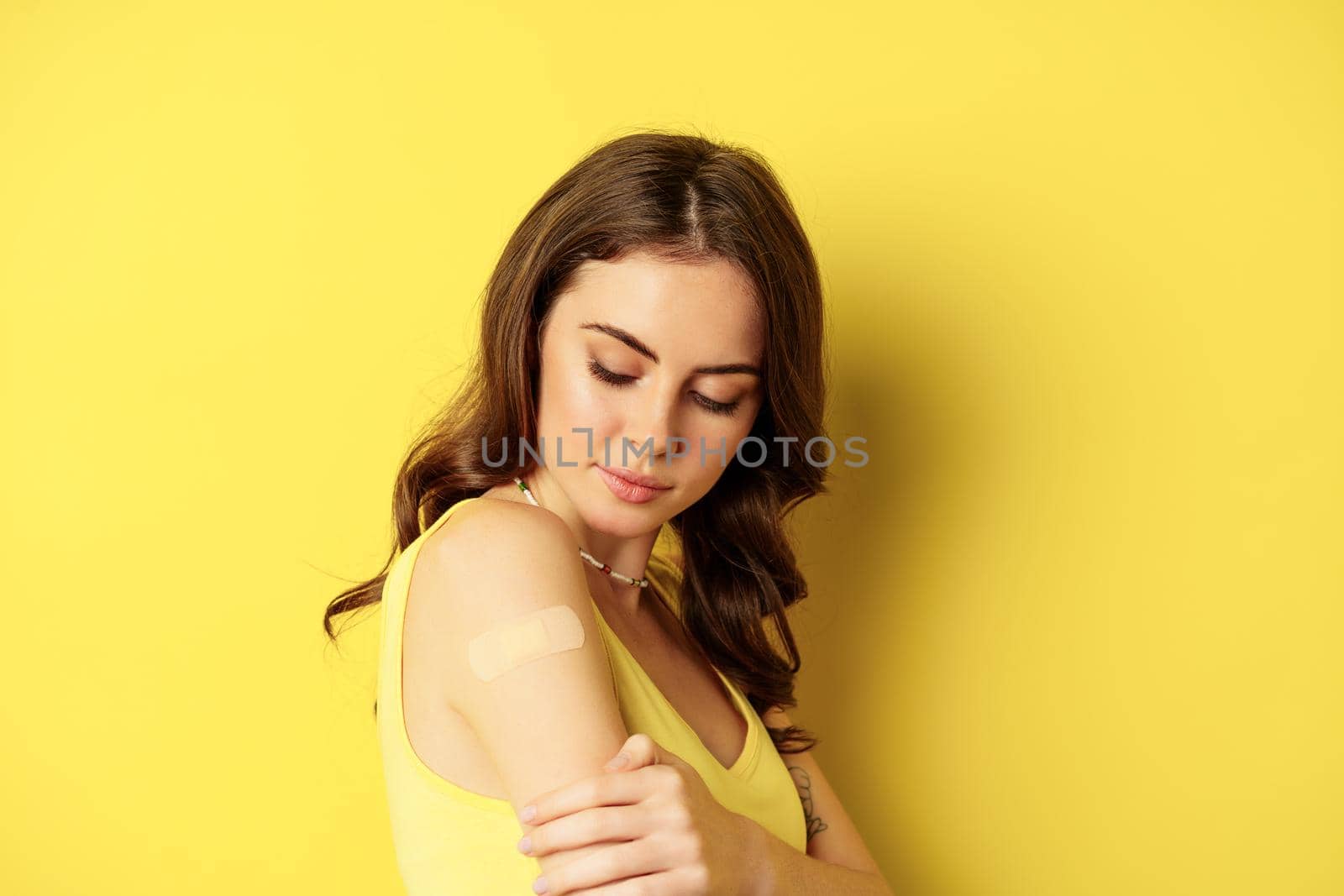 Covid-19 vaccination. Close up portrait of young millennial woman looking at patch on shoulder after vaccine from coronavirus shot, standing against yellow background.