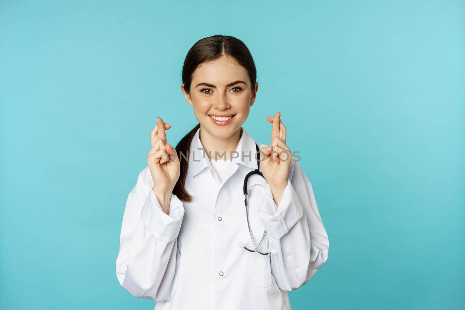 Hopeful woman doctor, lab worker cross fingers and smiling, praying, making wish, anticipating, standing over torquoise background.