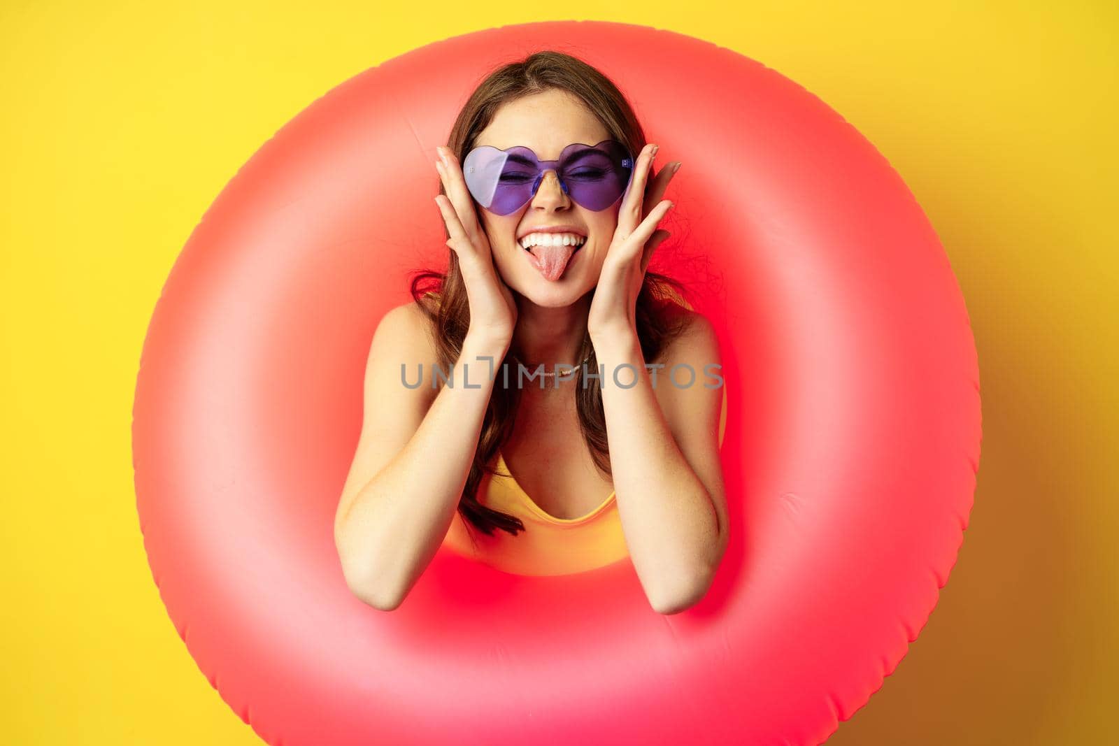 Close up portrait of enthusiastic young woman inside pink swimming ring, laughing and smiling, enjoying beach holiday, summer vacation, yellow background by Benzoix