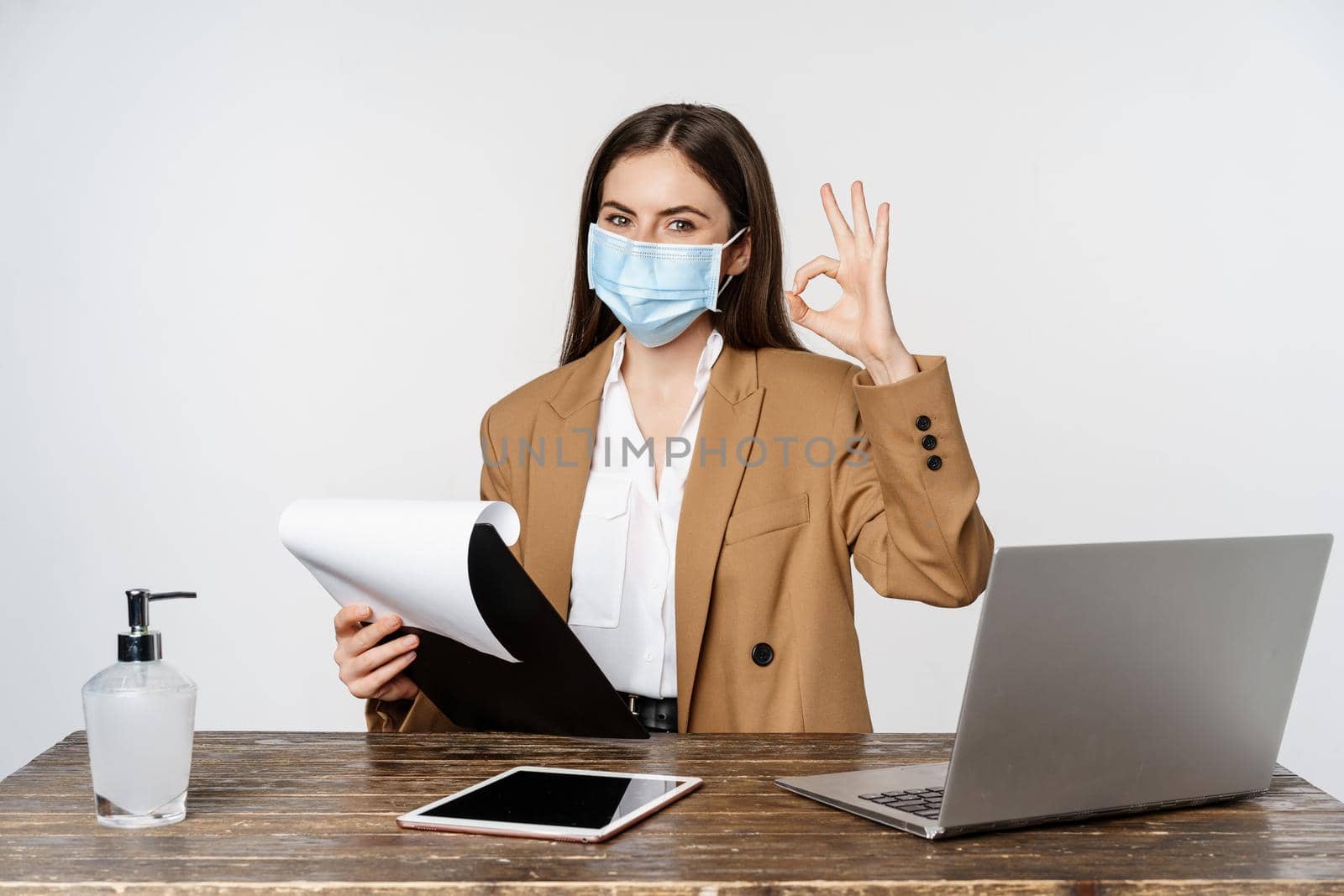 Woman working in office at table, holding clipboard with papers, showing okay sign, wearing medical face mask, white background. Copy space