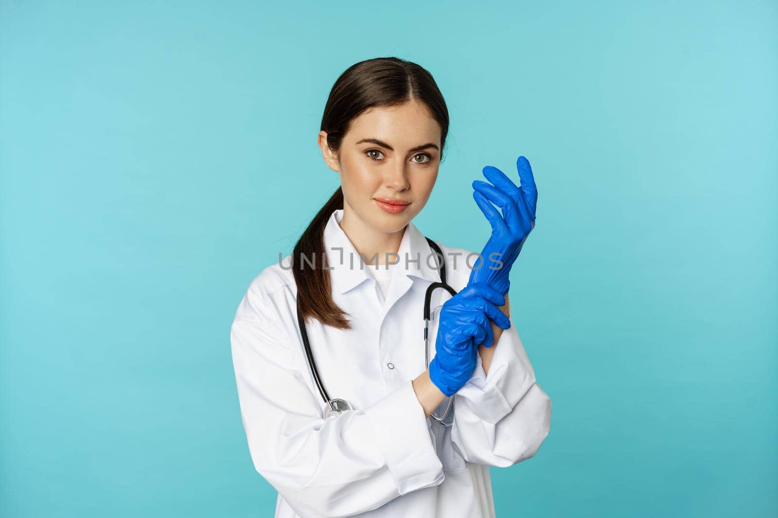 Young doctor, professional physician woman, put on gloves, smiling at camera, ready for medical checkup, standing in uniform over blue background.