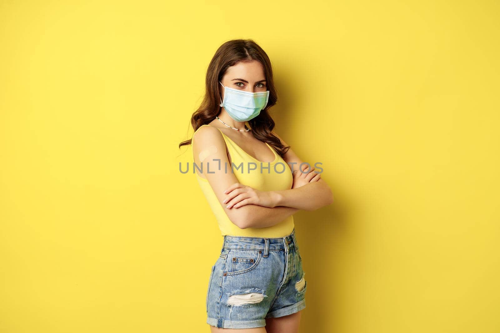 Covid-19 vaccination. Beautiful young woman in face medical mask got vaccinated during pandemic, standing over yellow background by Benzoix