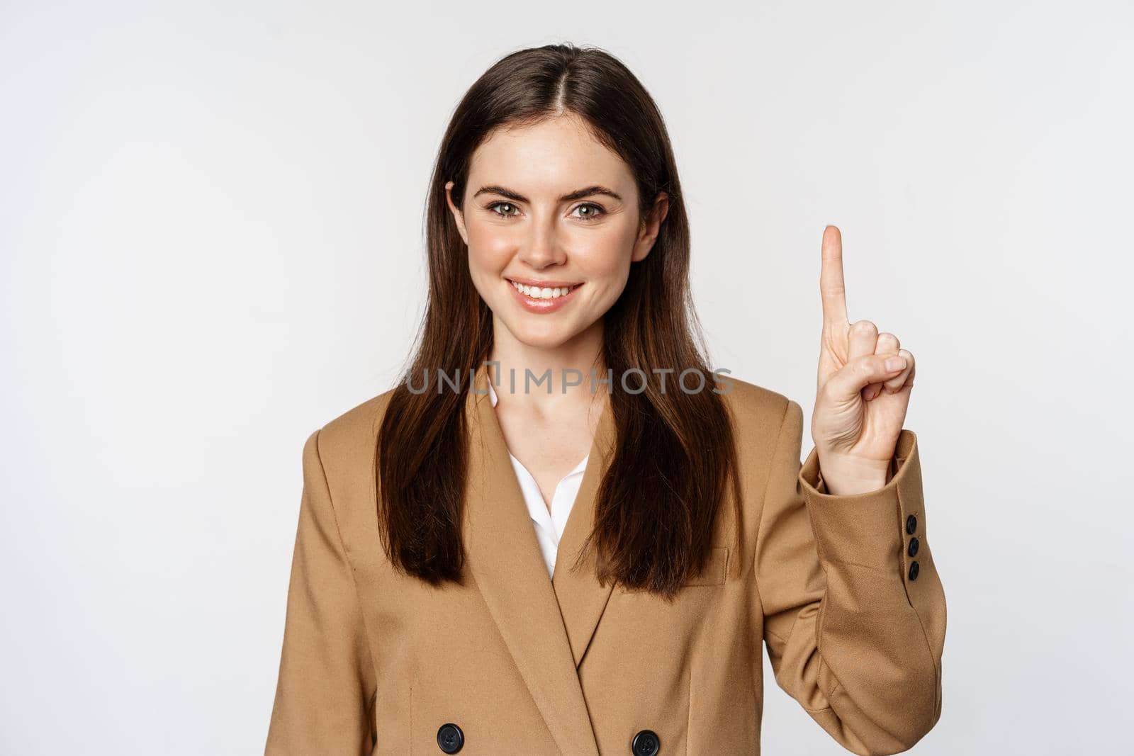 Portrait of corporate woman, saleswoman showing number one finger and smiling, standing in suit over white background.