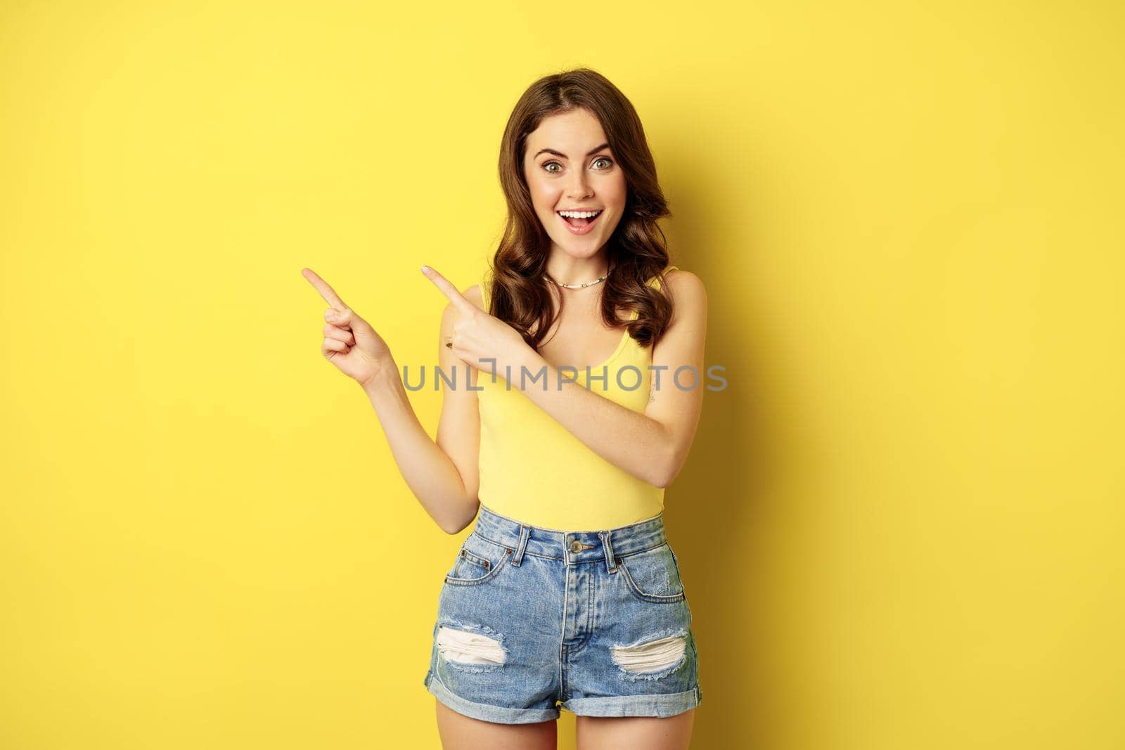 Portrait of stylish summer girl, brunette woman showing way, pointing fingers left, advertising, standing over yellow background by Benzoix