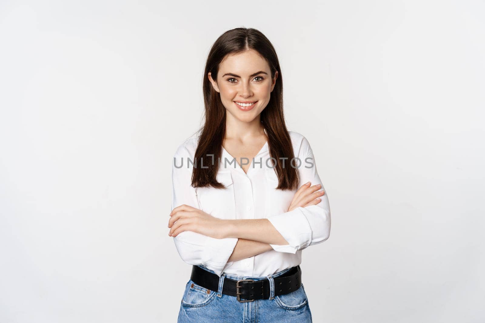 Smiling young businesswoman, female entrepreneur in white shirt, cross arms on chest like professional, standing over studio background.