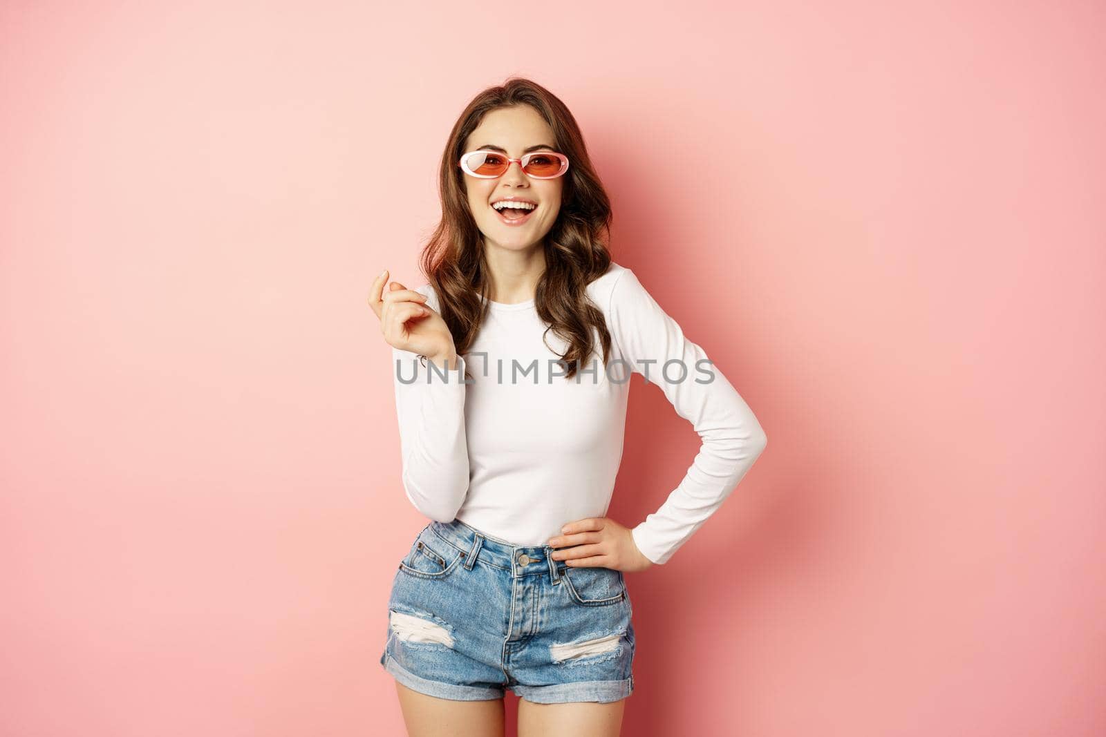 Portrait of stylish glam girl in sunglasses, laughing and smiling, standing over holiday pink background by Benzoix