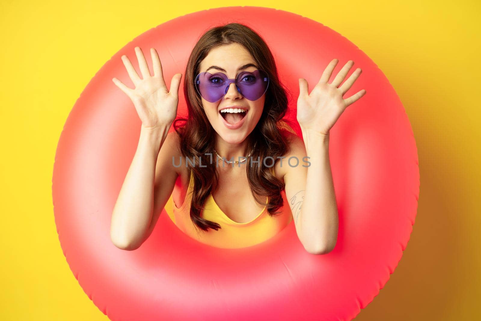 Close up portrait of enthusiastic young woman inside pink swimming ring, laughing and smiling, enjoying beach holiday, summer vacation, yellow background.
