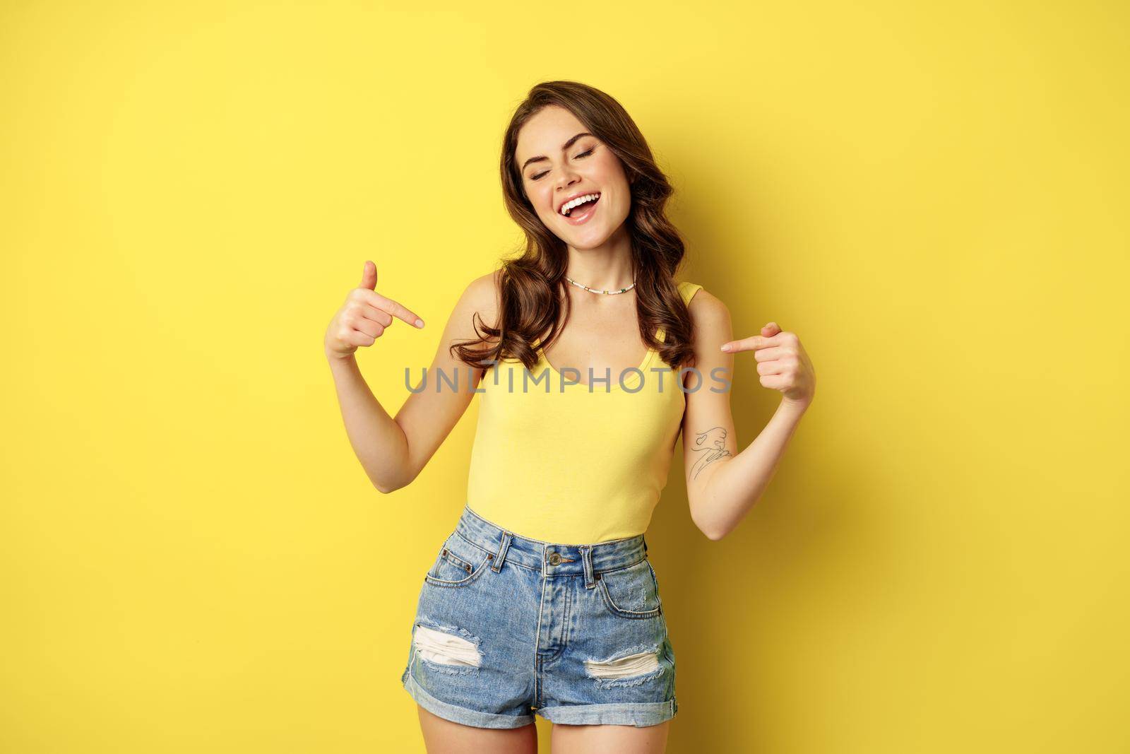 Happy attractive woman pointing fingers at herself, self-promoting, something personal, smiling and looking proud or confident, standing over yellow background.