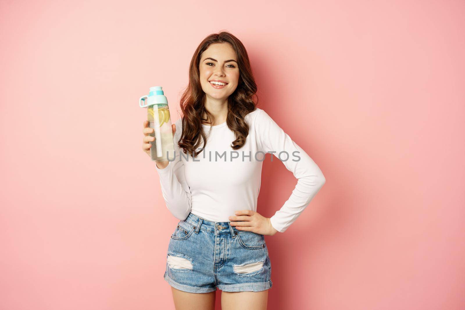 Healthy lifestyle. Happy smiling woman holding water bottle with lemon, vitamin c drink, looking joyful at camera, posing against pink background.
