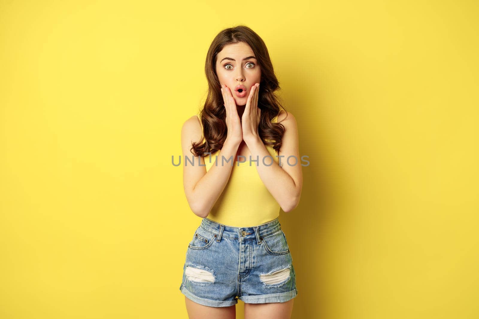 Portrait of brunette woman reacting surprised to advertsement, gasping and saying wow, wearing summer clothes, standing over yellow background.