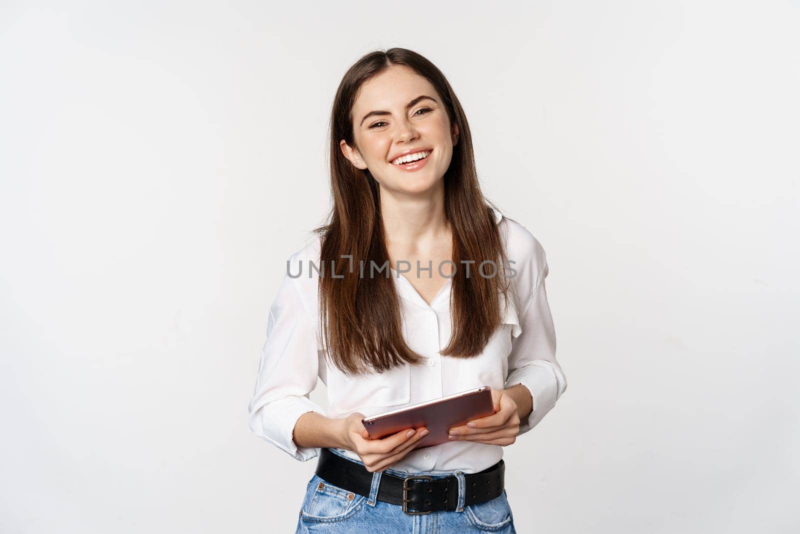Smiling modern woman standing with digital tablet, laughing and looking happy, working, posing against white studio background.