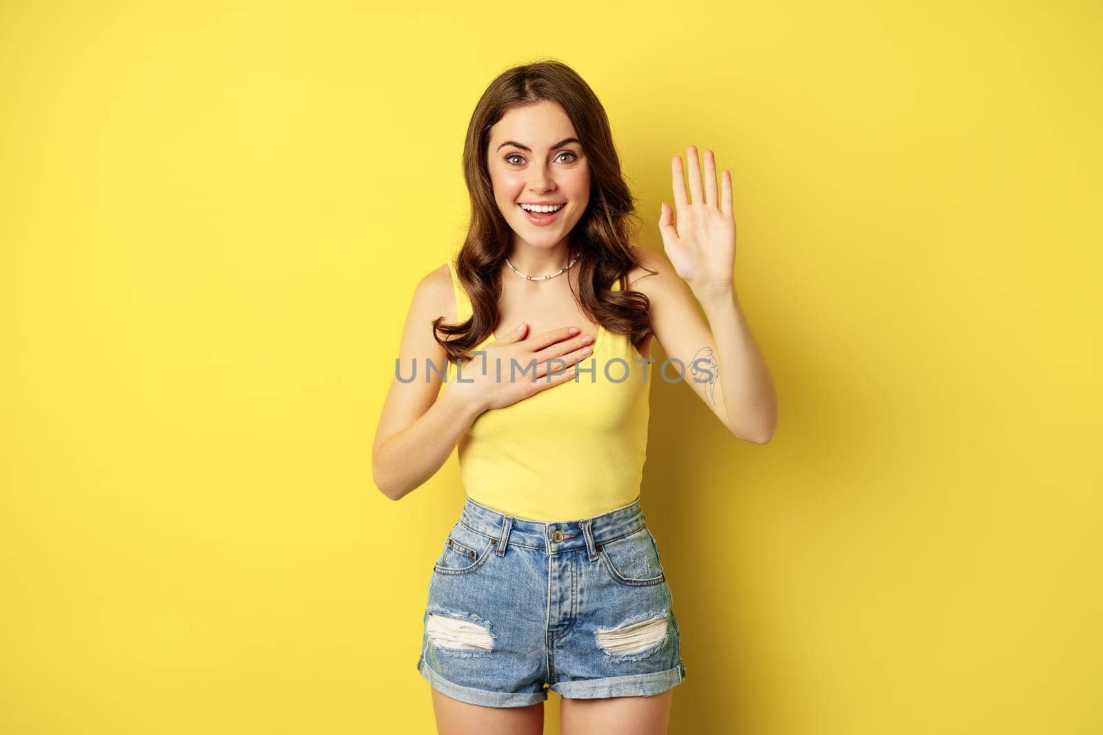 Enthusiastic girl raising hand, promise, introduce herself, say her name, standing over yellow background, smiling and looking excited by Benzoix