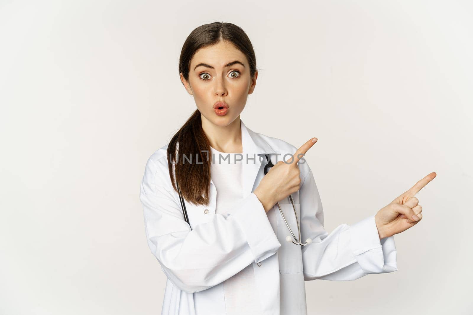 Woman doctor, physician in medical uniform, pointing fingers right, showing banner advertisement, smiling amazed, checking out promo discount, white background.