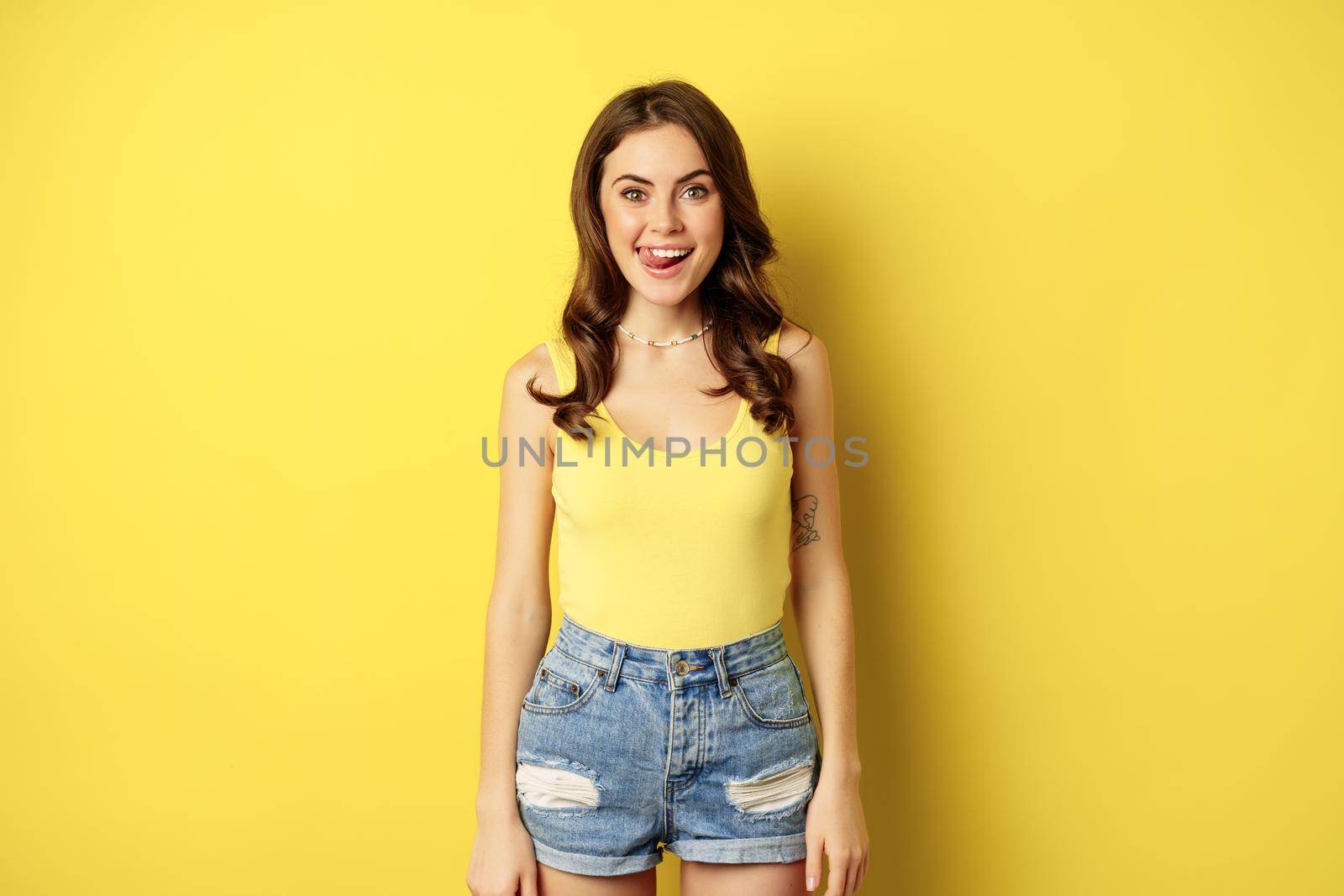 Stylish young brunette woman showing her white healthy teeth, smiling and showing tongue, winking enthusiastic, standing over yellow background by Benzoix