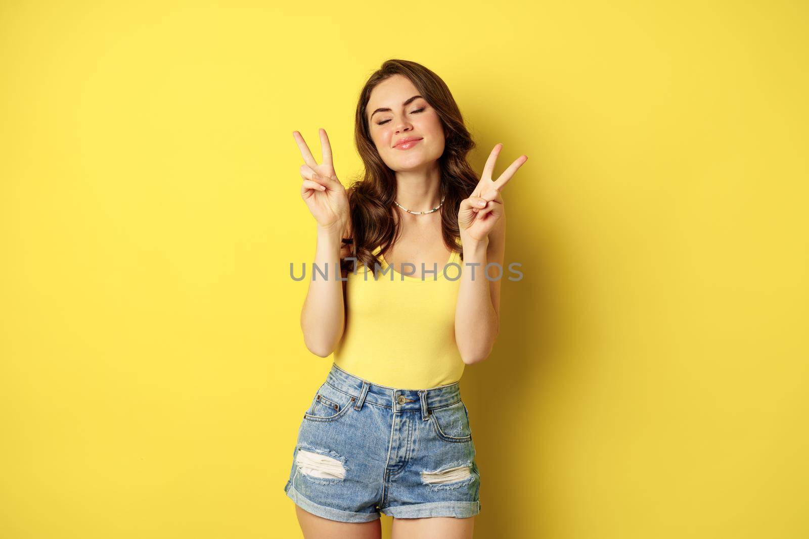 Positive girl, female model showing peace, v-sign gesture and smiling, standing in tank top and denim shorts, yellow background by Benzoix