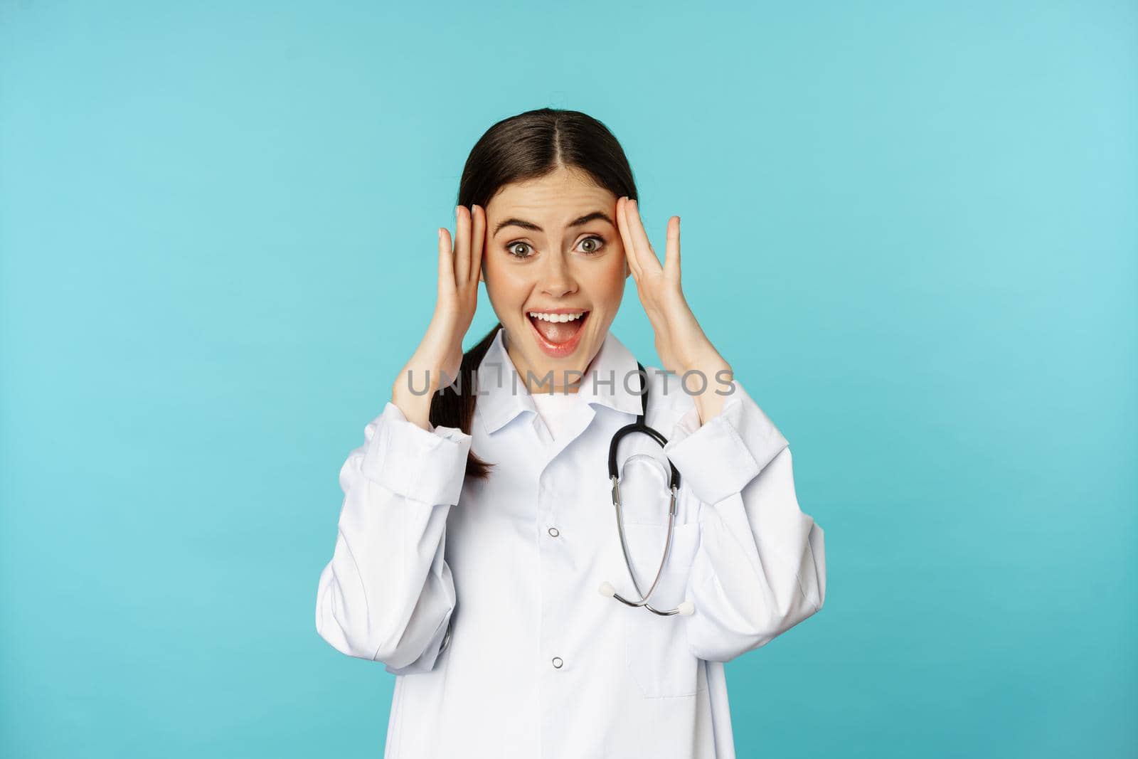 Portrait of surprised and excited smiling doctor woman, holding hands on head, reaction to mindblowing awesome news, standing over blue background.
