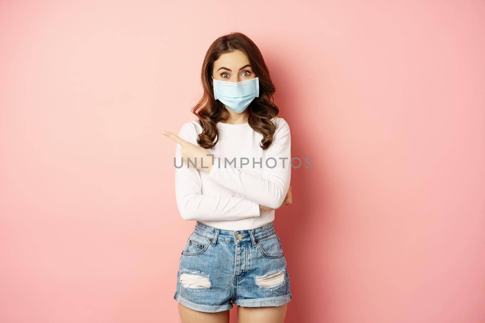 Beautiful stylish woman in medical face mask from covid, pointing finger at upper left corner, showing advertisement, standing over pink background.