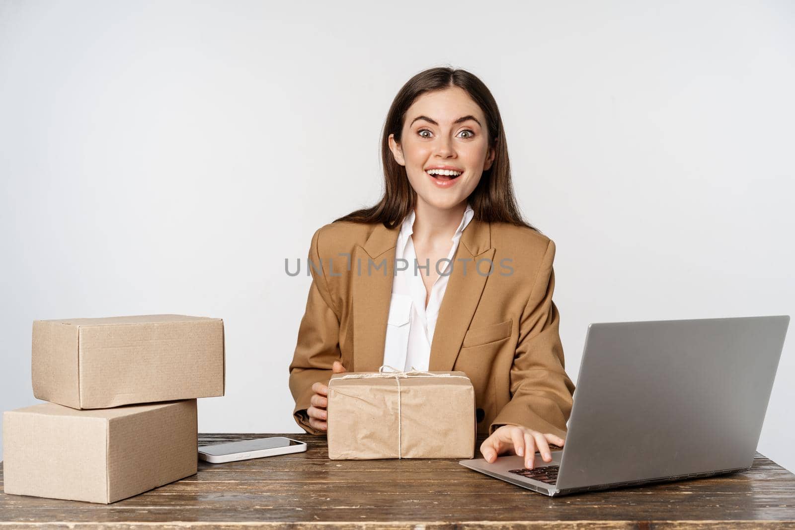 Smiling happy businesswoman working in her office with packages delivery, process orders on her laptop website, sending goods to clients, white background.