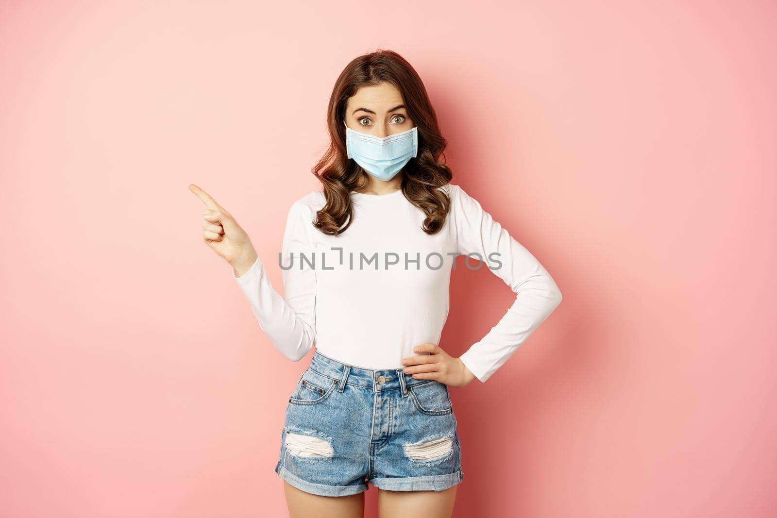 Beautiful stylish woman in medical face mask from covid, pointing finger at upper left corner, showing advertisement, standing over pink background by Benzoix