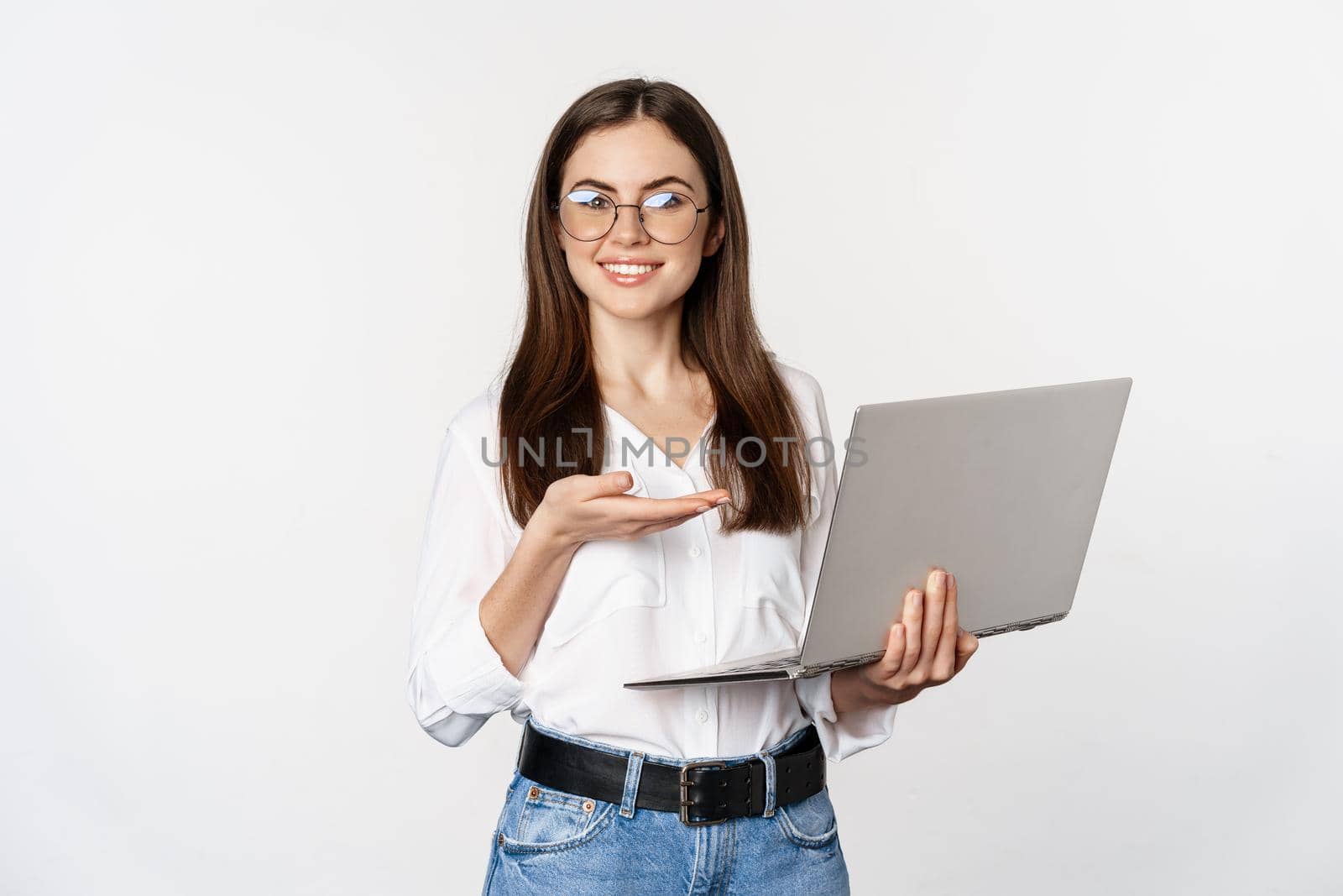 Portrait of woman in glasses holding laptop, pointing at screen, showing her work on computer, standing over white background.