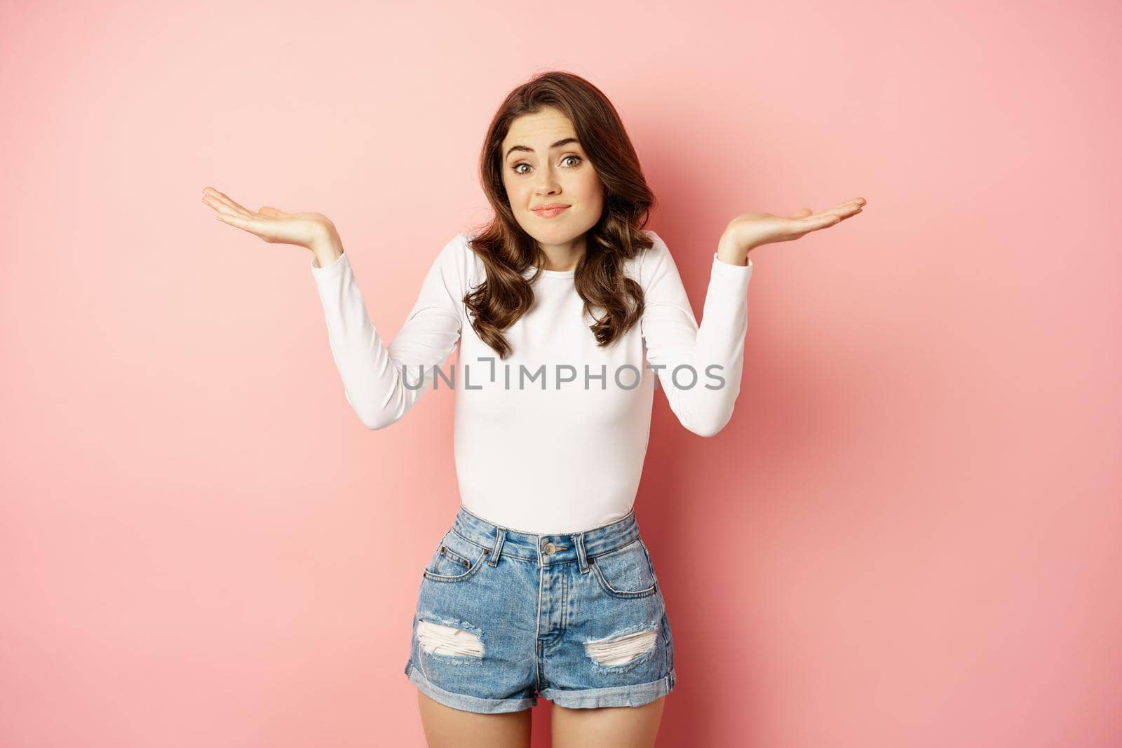 I dont know. Confused cute young woman smiling awkward, shrugging shoulders clueless, has no idea, standing over pink background.