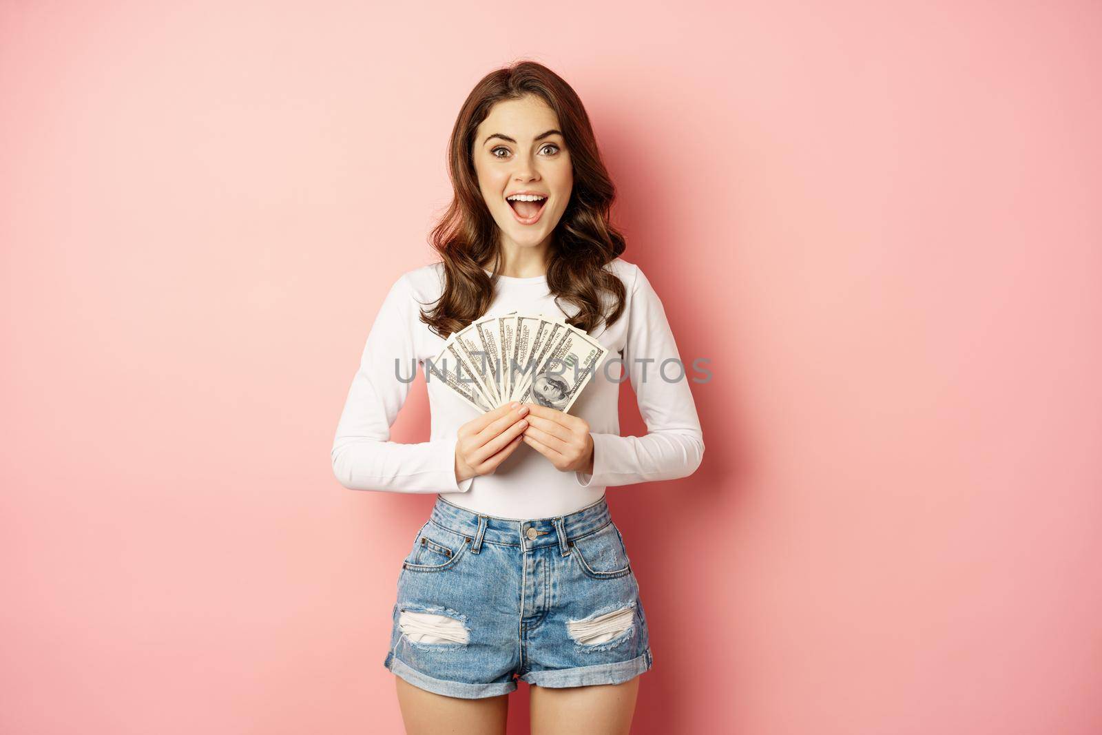 Loans and microcredit. Smiling beautiful girl showing money, cash in hands and looking enthusiastic, standing over pink background.