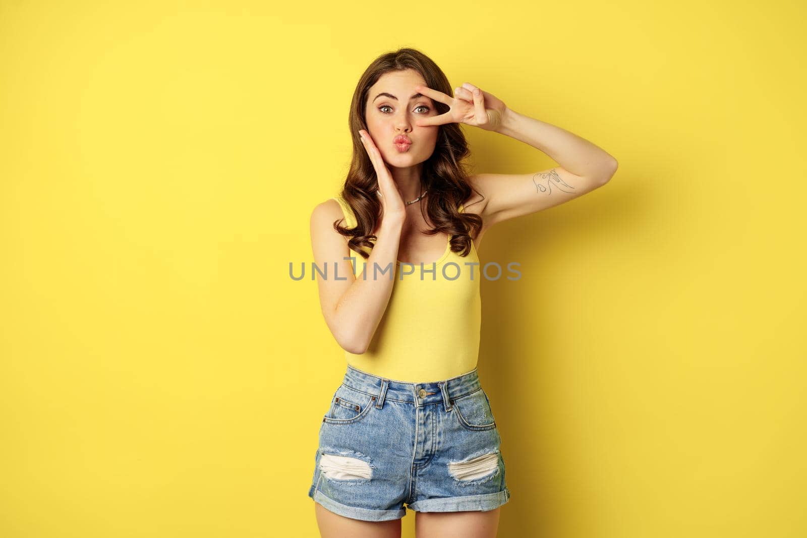 Beautiful glamour girl in summer outfit, showing peace sign, kissing face, standing against yellow background.