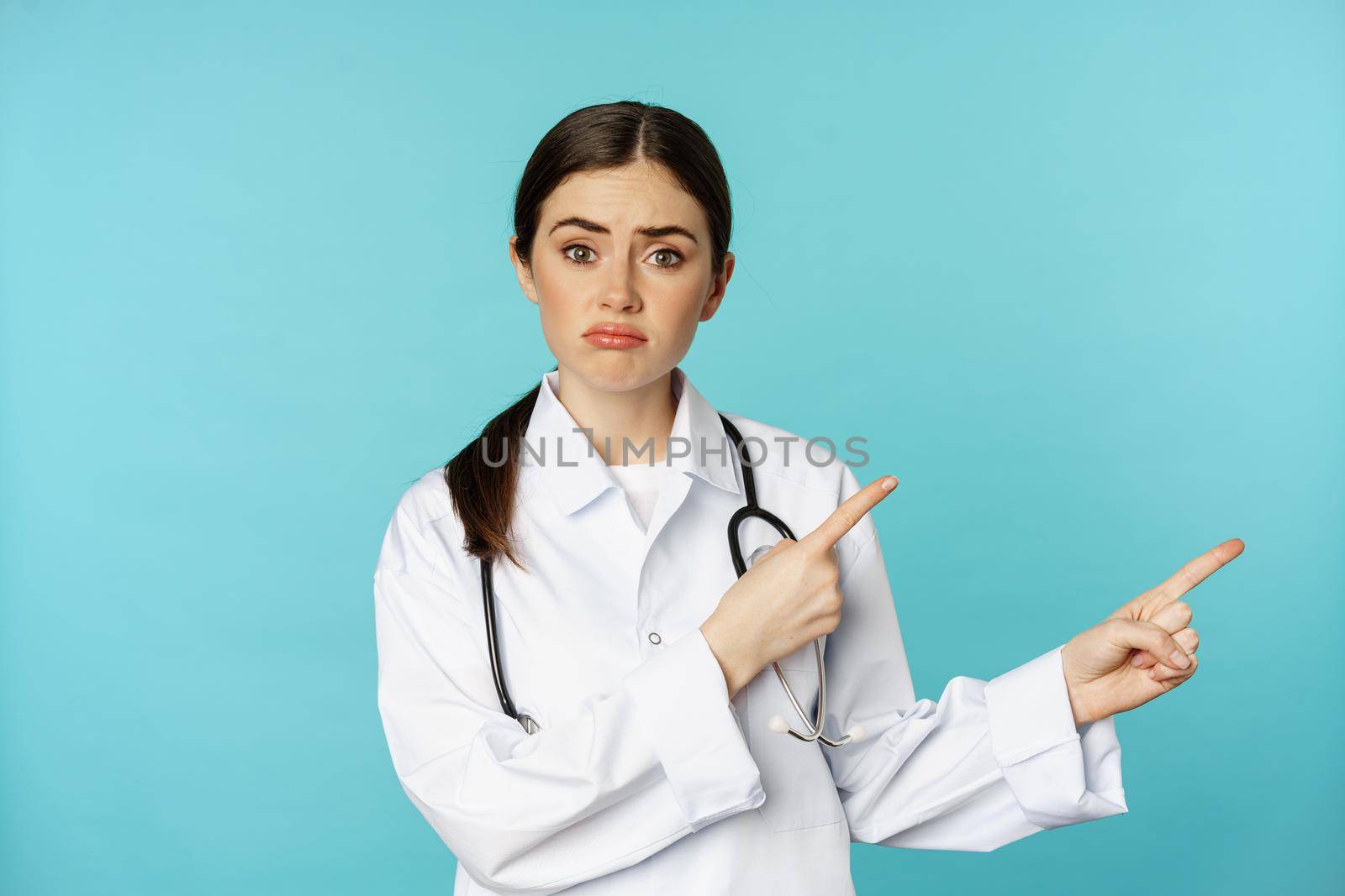 Portrait of disappointed doctor, woman medical worker pointing fingers right and looking sad, regret, gloomy face expression, blue background.