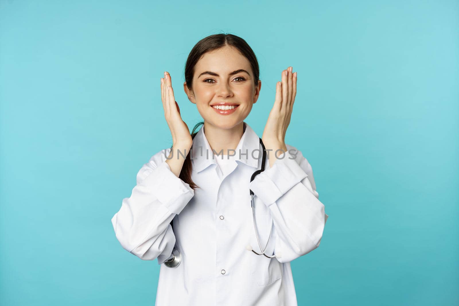 Happy smiling woman doctor, therapist open eyes and looking surprised at camera, standing in white coat over blue background.