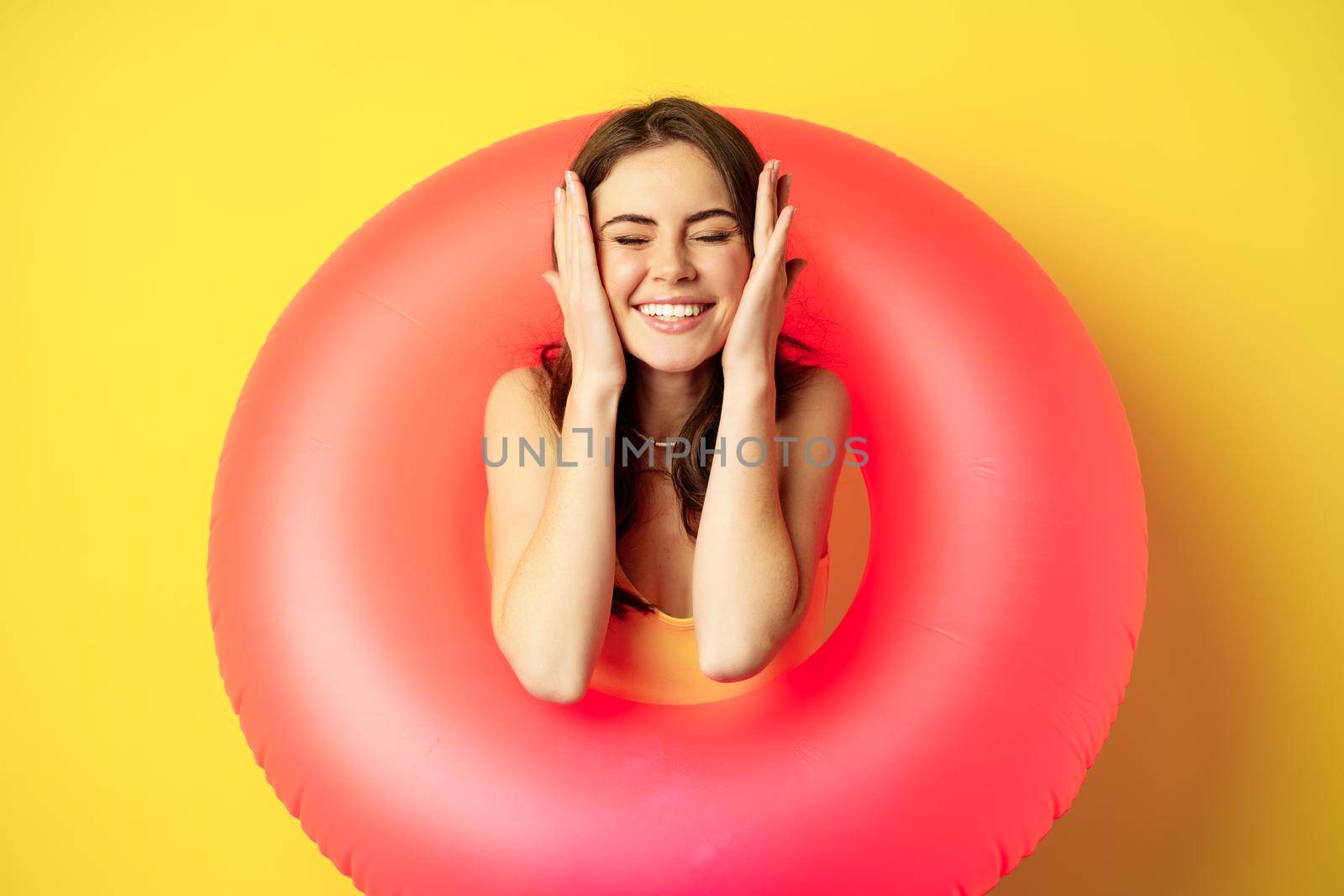Happy beautiful young woman relaxing on summer vacation, wearing pink swim ring for beach holiday, standing against yellow background.
