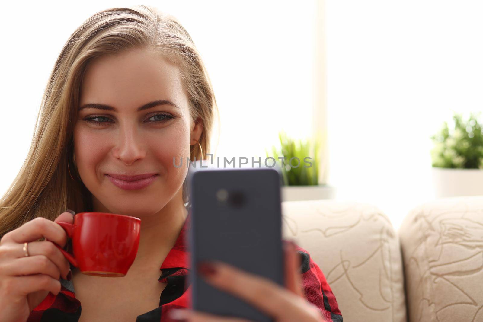 Young beautiful woman drinks coffee and looks into smartphone. Positive online communication concept