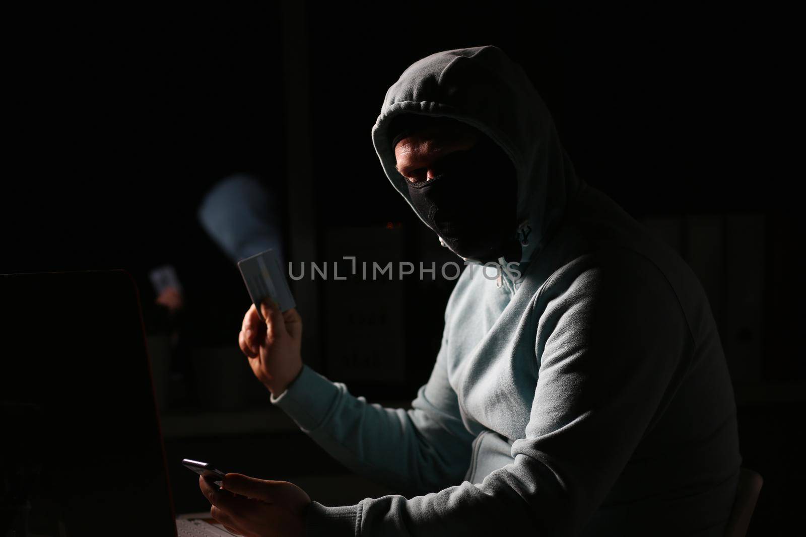 Hacker in dark jacket looking at laptop screen in dark room is holding bank card by kuprevich