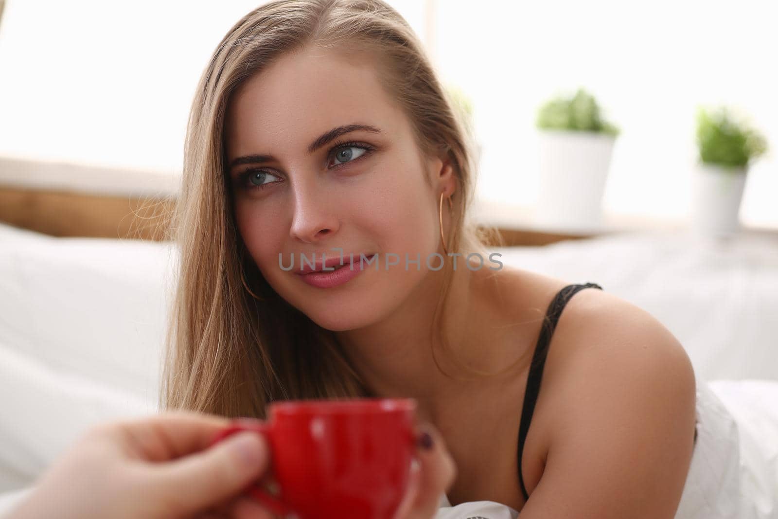 Young smiling blonde is drinking cup of tea with coffee early in morning while lying in bed. Morning awakening and water concept