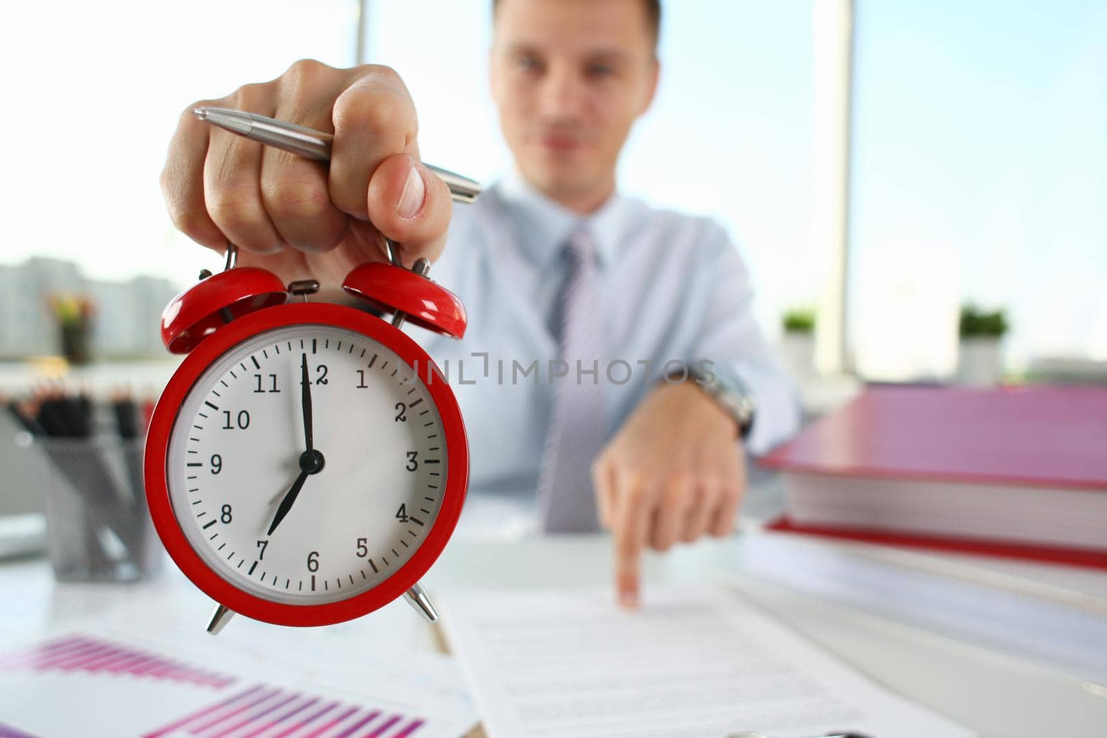 Man hand on red alarm clock stands at desk in office showing seven o'clock in morning or in evening AM PM. Overtime work deadline concept