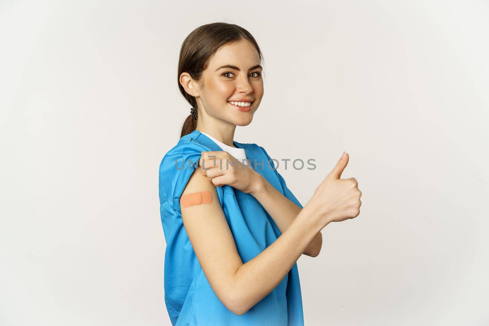 Smiling nurse, doctor in scrubs, medical worker showing her vaccinated shoulder with patch, thumbs up, recommending vaccinate from coronavirus, white background.