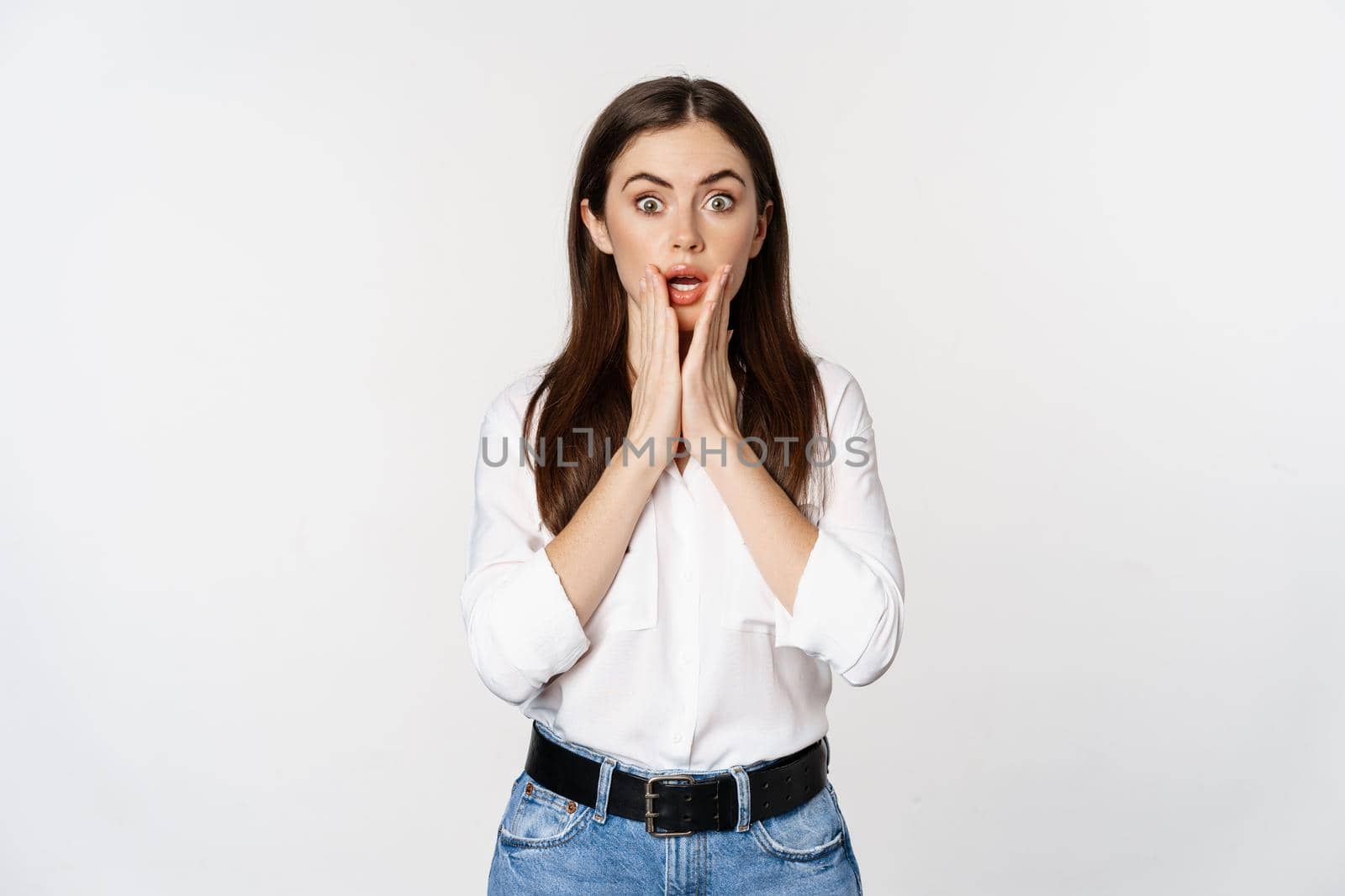 Surprised brunette woman looking amazed, seeing smth awesome, standing in blouse and jeans over white background.