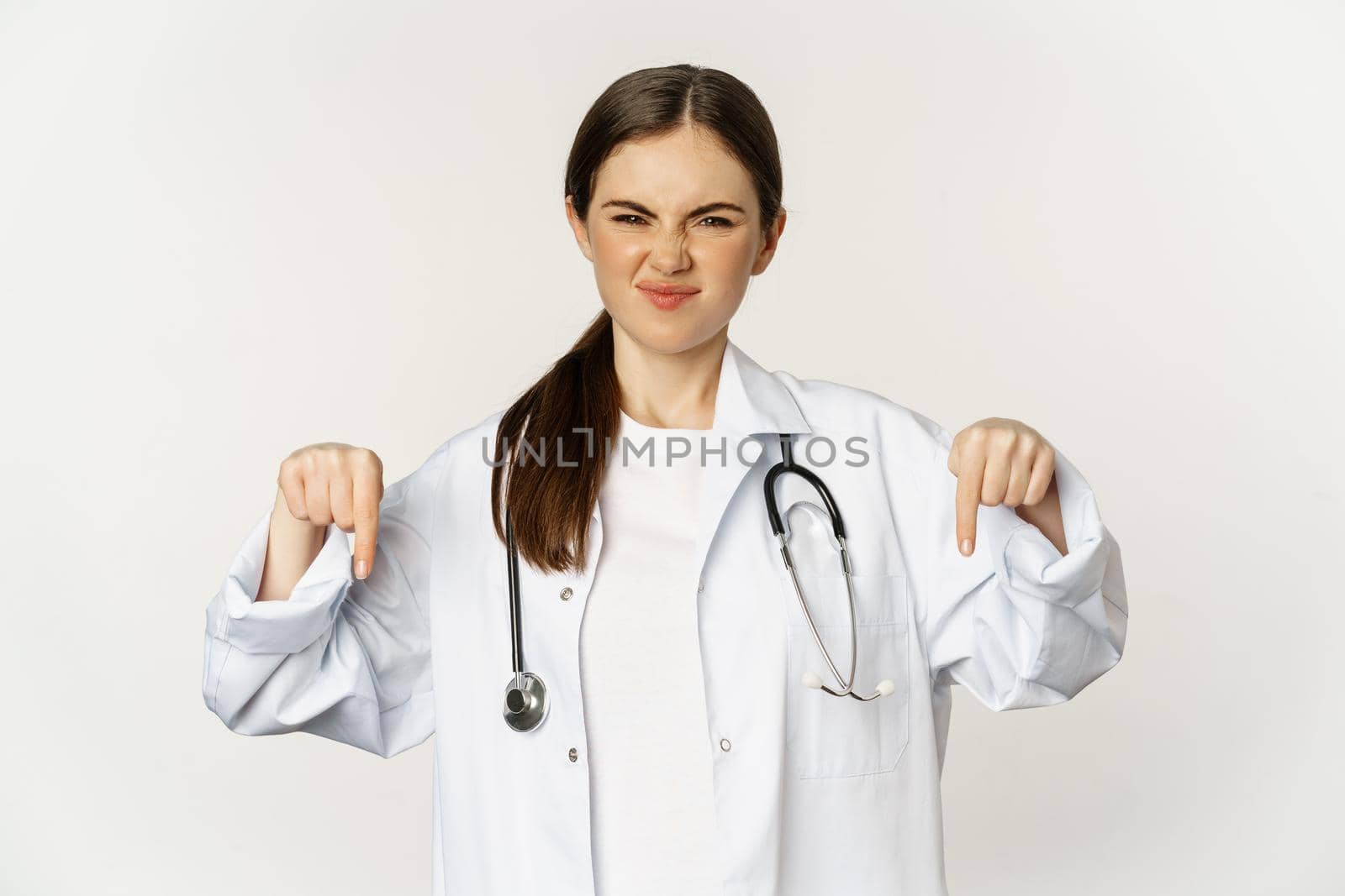 Disappointed young doctor, female physician pointing fingers down and grimacing with dislike, disapprove smth, standing over white background.
