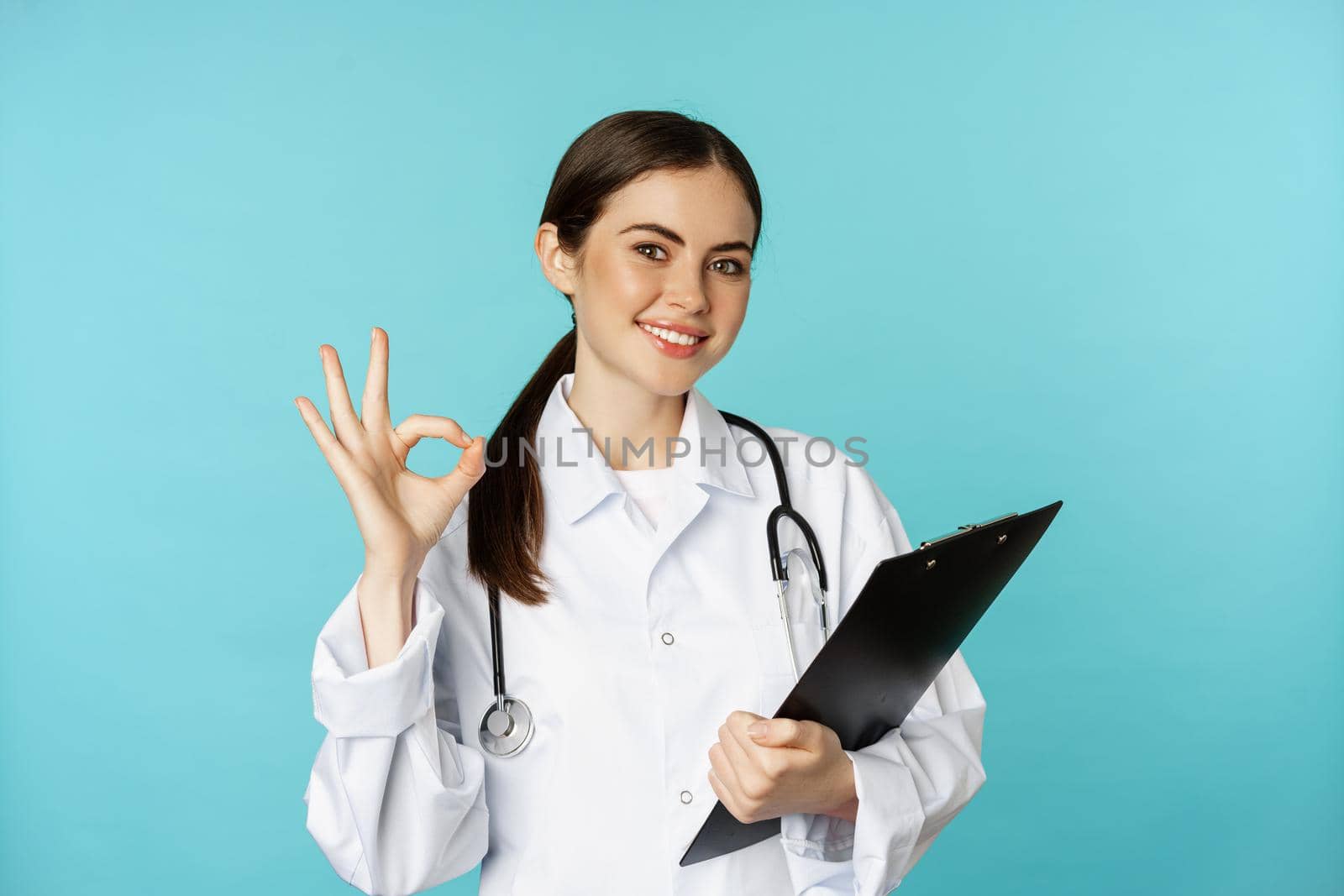 Confident smiling doctor woman, physician showing okay, holding clipboard, appointment in hospital, standing over torquoise background.