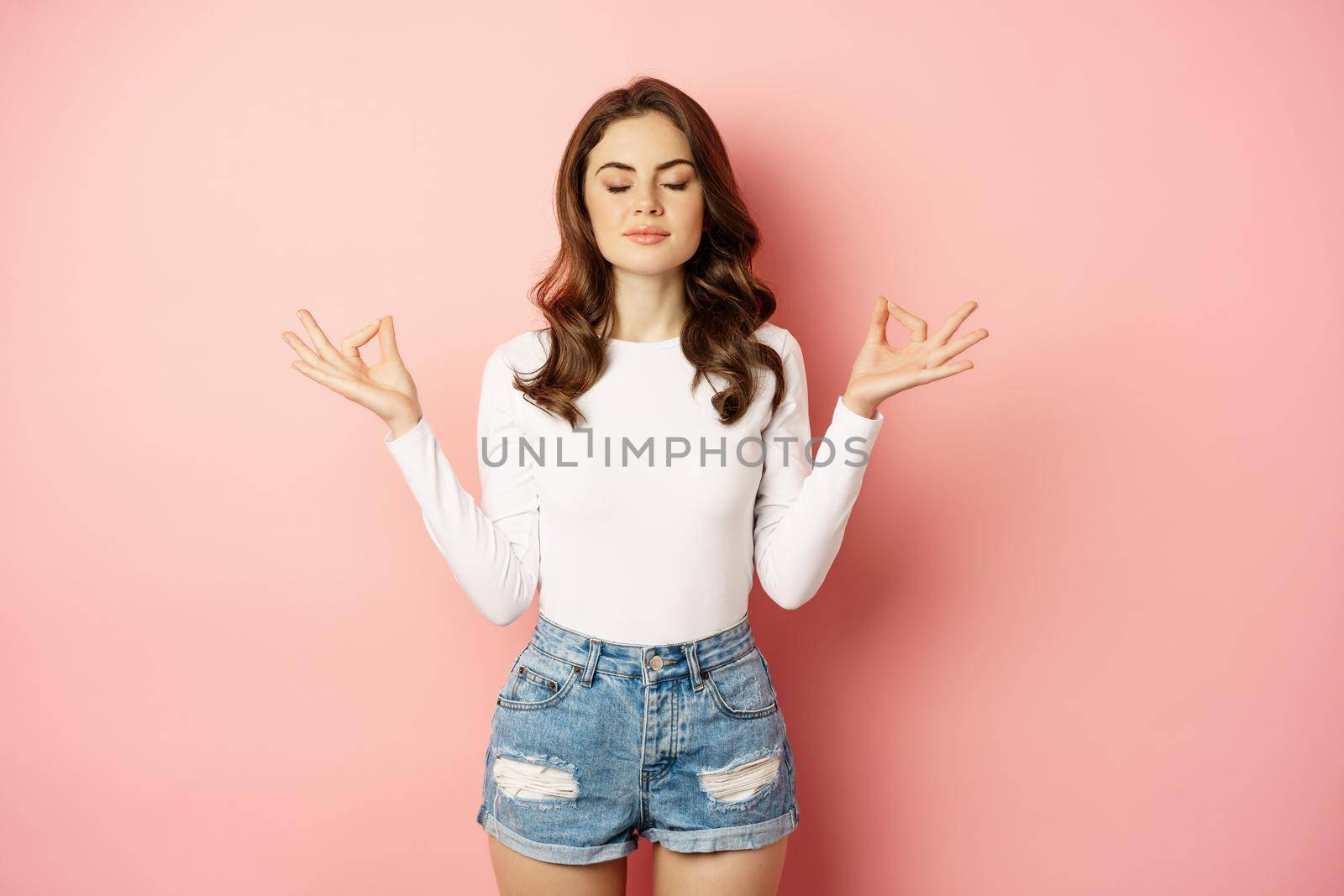 Relaxed mindful young woman meditating, keep calm, relaxing during yoga, close eyes and fold fingers in mudra gesture, pink background.