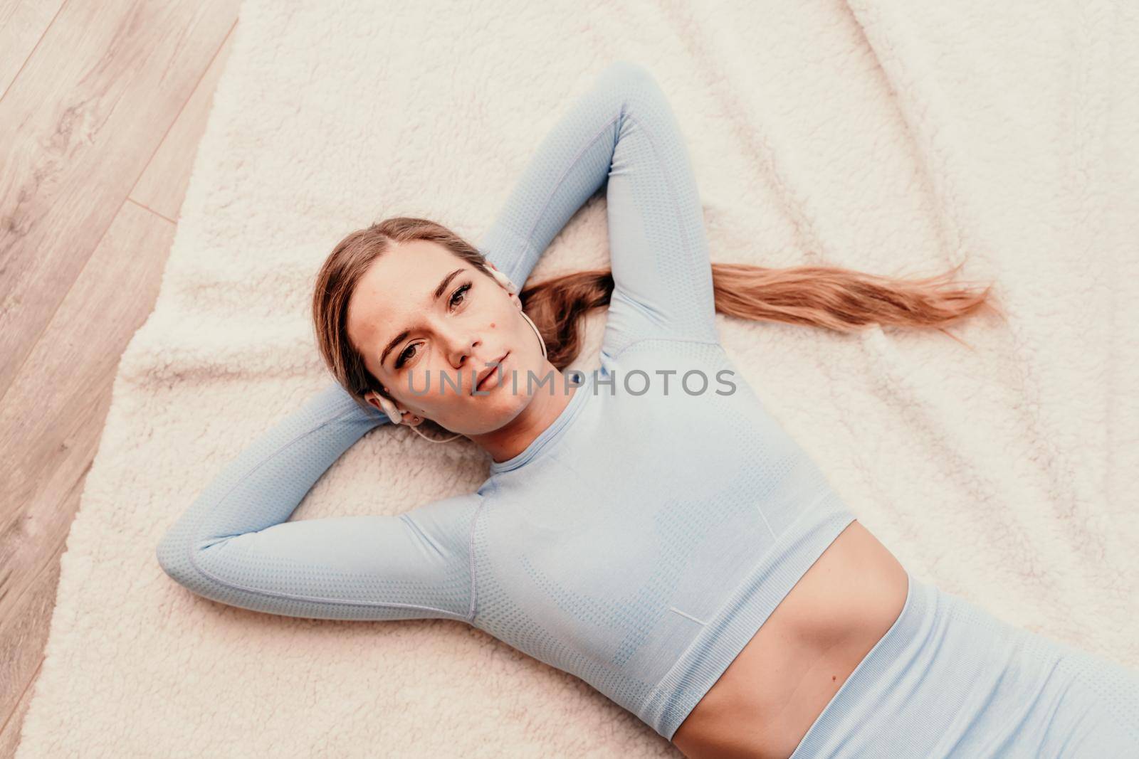 Top view portrait of relaxed woman listening to music with headphones lying on carpet at home. She wears a blue suit with long hair pulled back into a ponytail